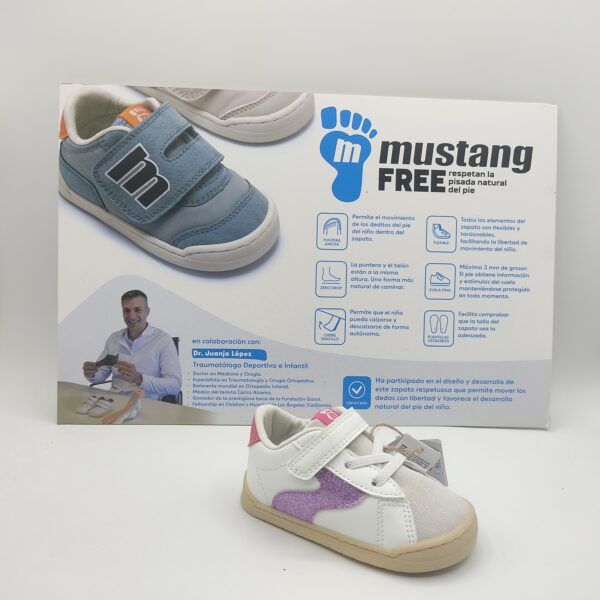 Zapatillas deportivas respetuosas para niñas de la marca Mtng, modelo 49304 en color blanco con detalles laterales y en el talón en color fuchsia. Suela blanca cosida. Parte delantera en serraje gris. Cierre con cordón elástico y velcro.